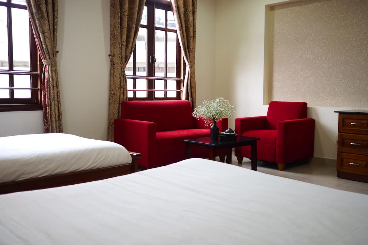 A hotel room with a large bed, two red armchairs, a small table with a vase, and a wooden dresser.