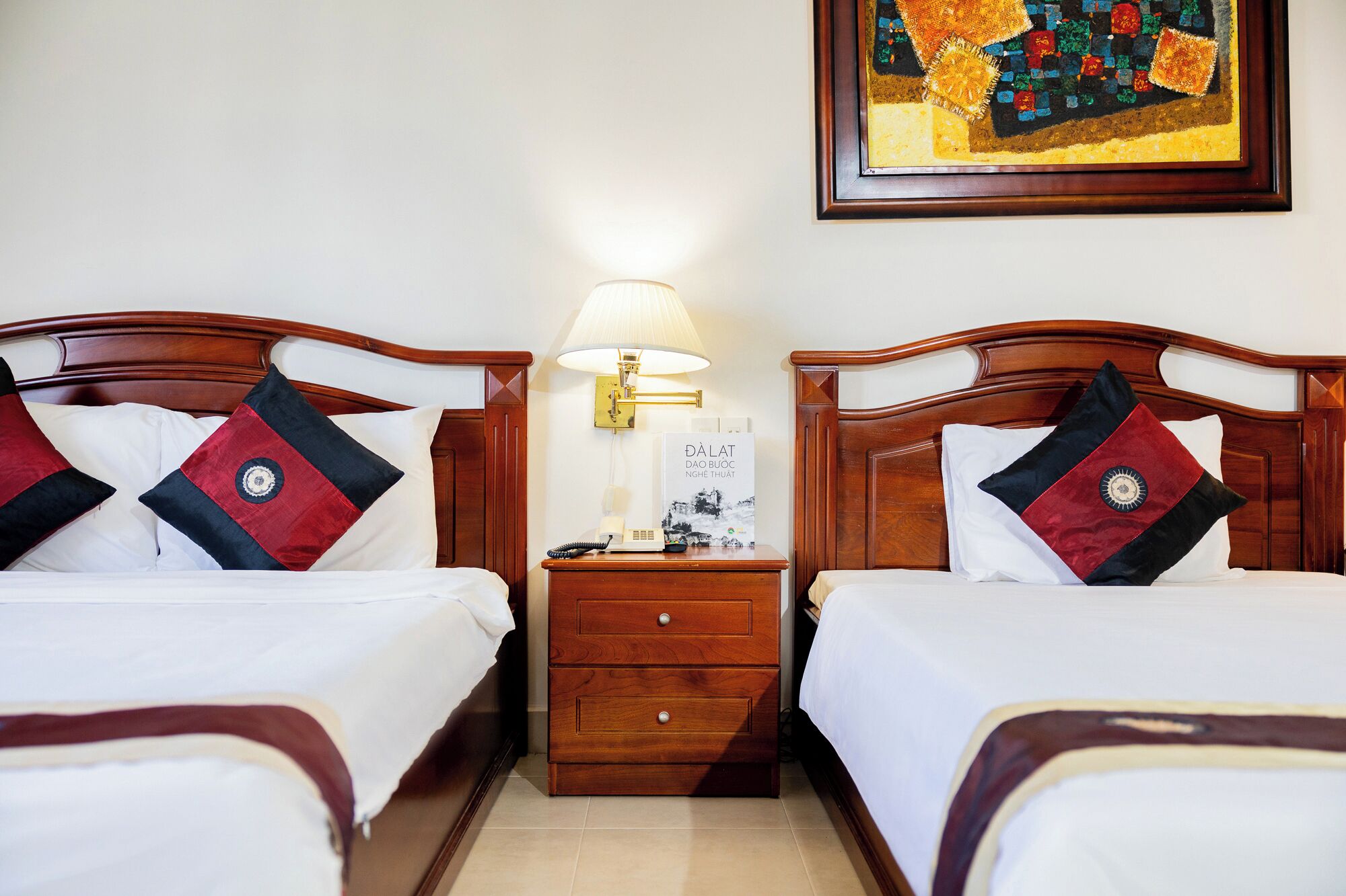 Een hotelkamer met twee houten bedden, elk voorzien van een witte sprei en decoratieve kussens. Er is een nachtkastje met een lamp, een klein tafeltje met een telefoon en een boek, en een ingelijst schilderij aan de muur.