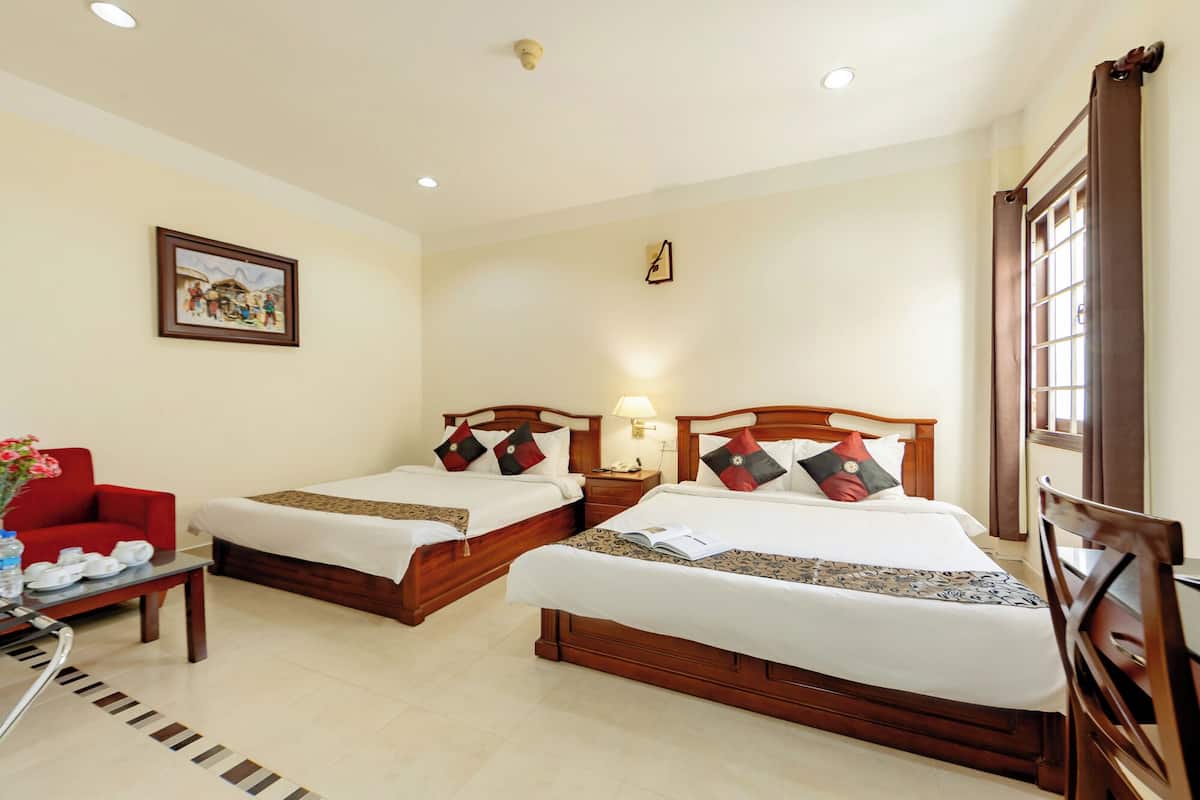 A hotel room with two beds, a red armchair, a small table with a vase of flowers, and a painting on the wall.
