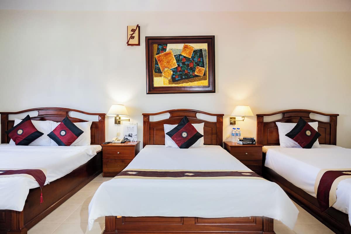 A hotel room with three beds, each with a wooden headboard and white bedding. There are bedside tables with lamps and a framed artwork above the beds.