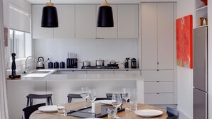 A modern kitchen with white cabinets, a central island, black pendant lights, and a dining area with chairs and a table set for four. - The Gallery - Central, Spa Pool, Sleeps 6 (Rotorua)