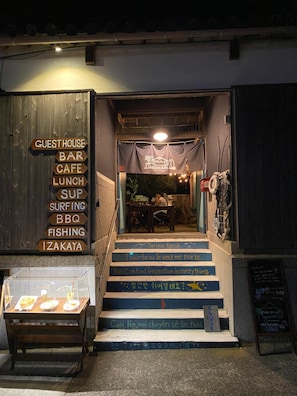 A wooden entrance with a sign listing amenities like guest house, bar, cafe, lunch, sup, surfing, bbq, fishing, and izakaya.