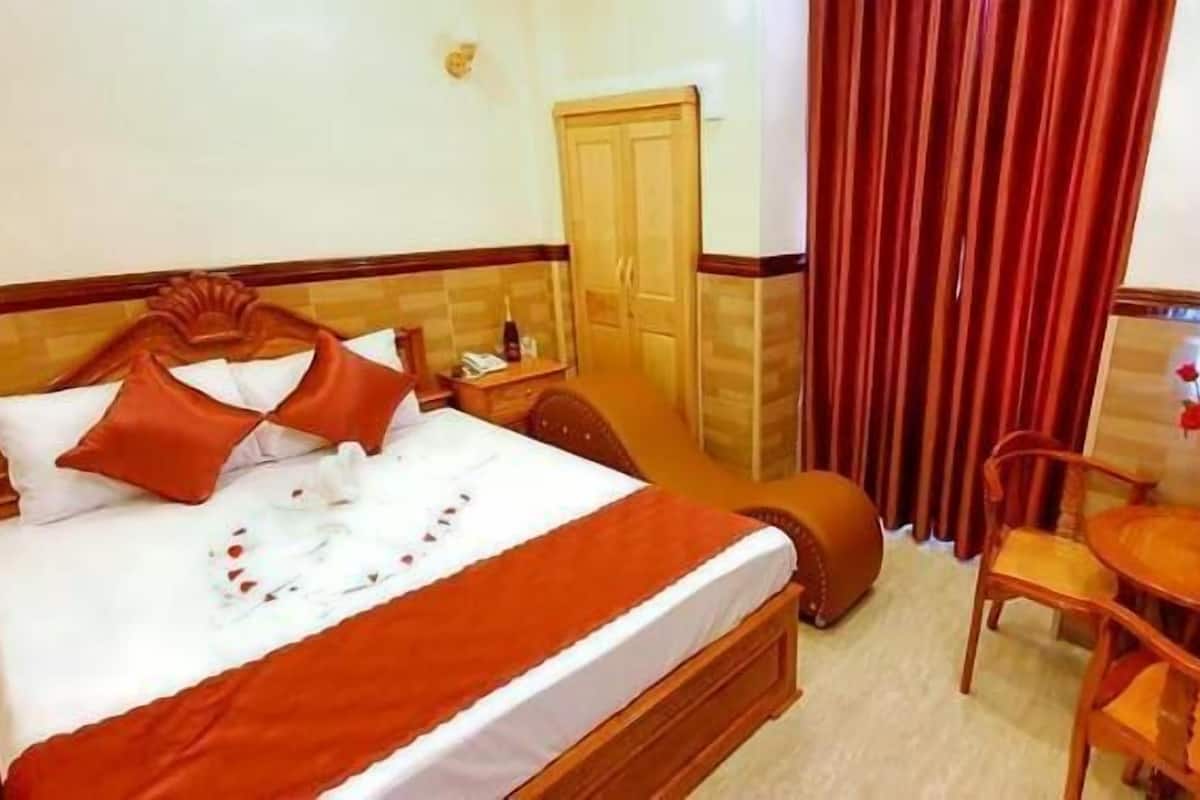 A hotel room with a bed, a wooden headboard, a desk with a chair, a small table with a vase of flowers, and a door with curtains.