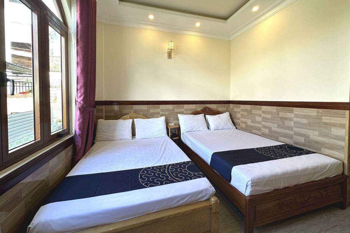 A hotel room with two beds, a window with curtains, a wall-mounted light fixture, and a view of the outside.