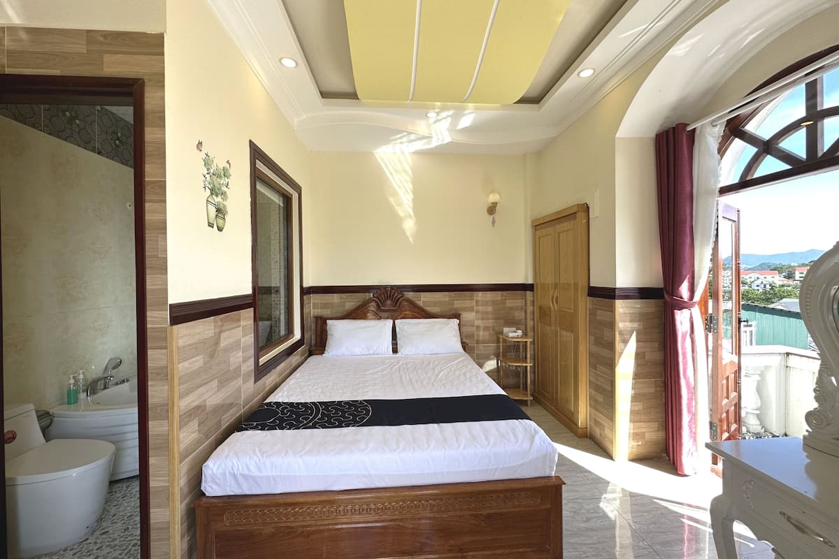 A hotel room with a large bed, a wooden wardrobe, a bathroom with a bathtub, and a view of the outside.