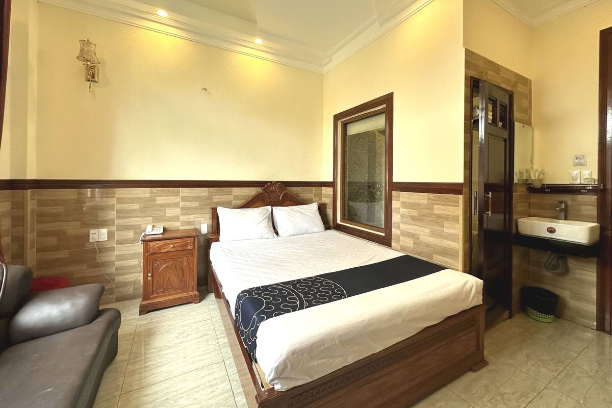 A hotel room with a bed, a wooden nightstand, a chair, a bathroom with a sink and mirror, and a tiled floor.