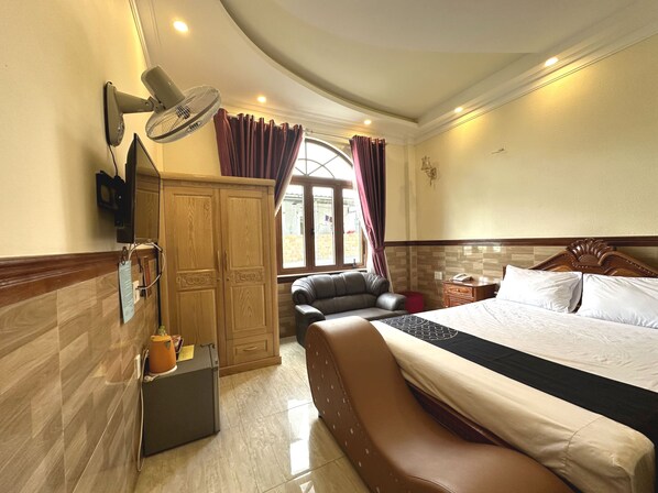 Habitación de hotel con una cama grande, un sofá de cuero, un armario de madera, un escritorio con lámpara y una ventana con cortinas.