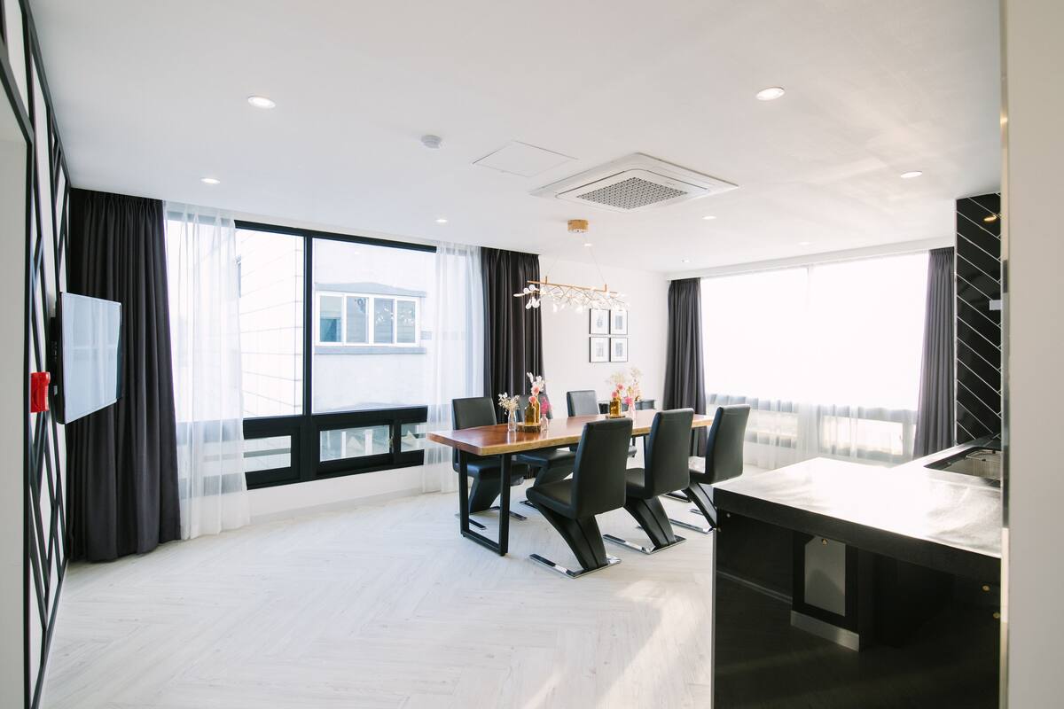 A modern hotel room with a large window, a dining area with a table and chairs, and a flat-screen TV mounted on the wall.