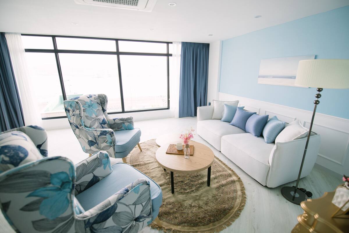 A modern living room with a large window, a white sofa with blue cushions, a floral armchair, and a round coffee table.