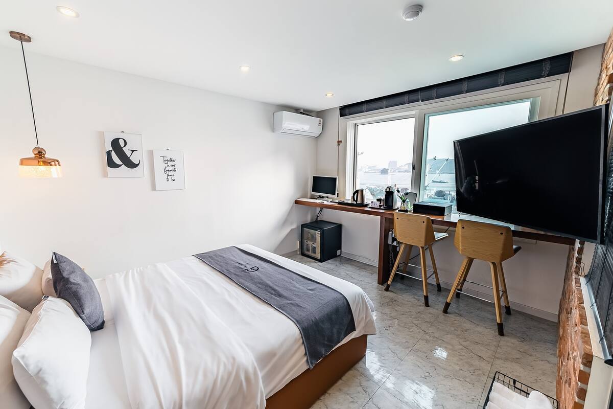 A modern hotel room with a bed, desk, TV, and large windows.