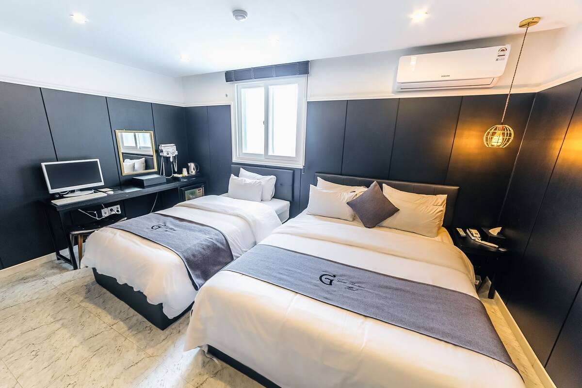 A modern hotel room with two beds, a desk with a computer, a mirror, and an air conditioning unit.