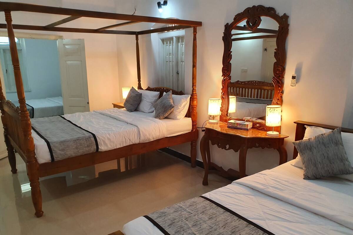 A bedroom with two beds, a wooden canopy bed, a nightstand with a lamp, a mirror, and a bedside table with books.