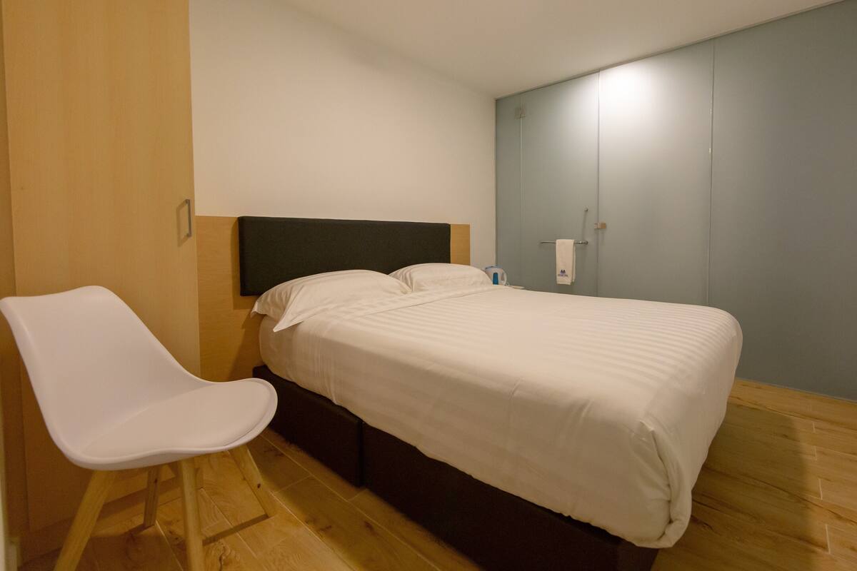 Un lit simple avec une tête de lit en bois, une chaise blanche et un interrupteur sur le mur.