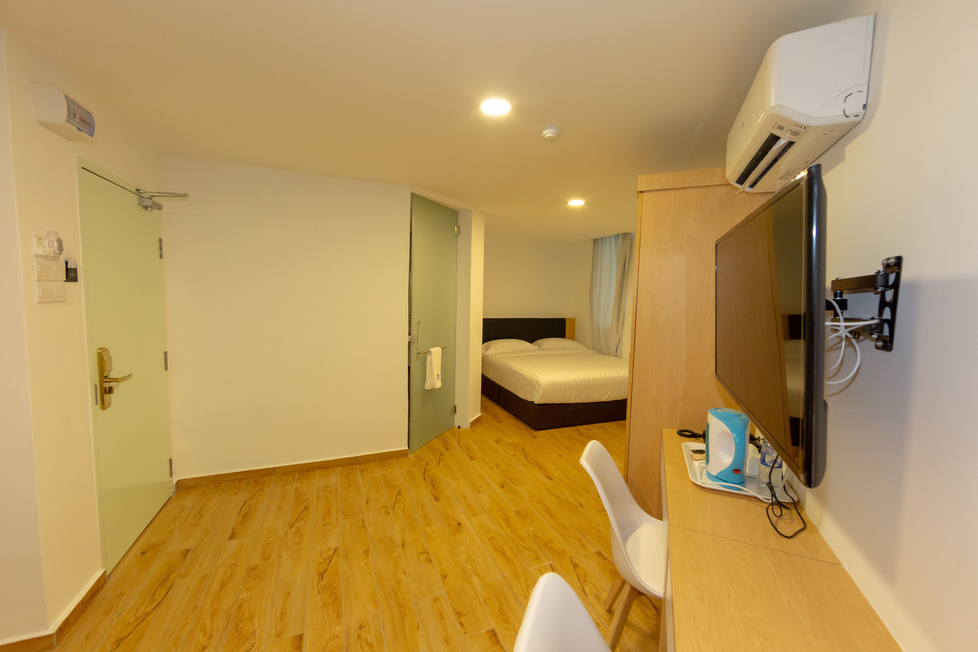 A hotel room with a wooden floor, a bed, a desk with a chair, a TV, and an air conditioning unit.