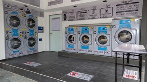 Een wasserette met meerdere wasmachines en drogers, waaronder modellen van 14kg en 16kg. Gebruiksaanwijzingen zijn op de muur aangebracht.