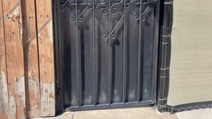 Una reja de metal negro con diseños decorativos, una cerca de madera en un lado y un muro de tela color beige en el otro.
