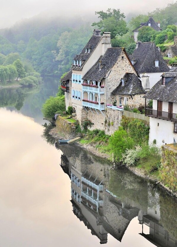 A riverside building with stone walls and multiple balconies, reflecting in the calm water.