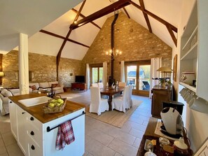 Una cocina amplia con pared de piedra, vigas de madera y zona de comedor con sillas blancas.