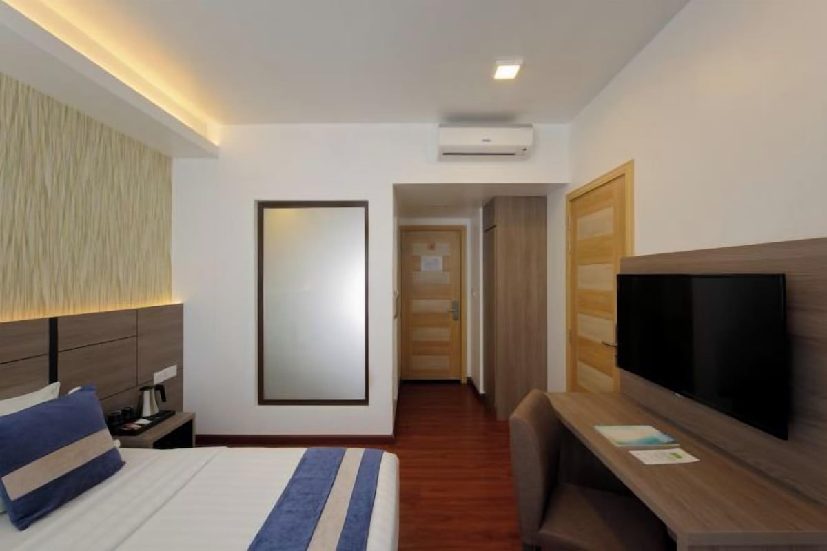 A hotel room with a bed, bedside table, lamp, TV, and a desk with a chair.