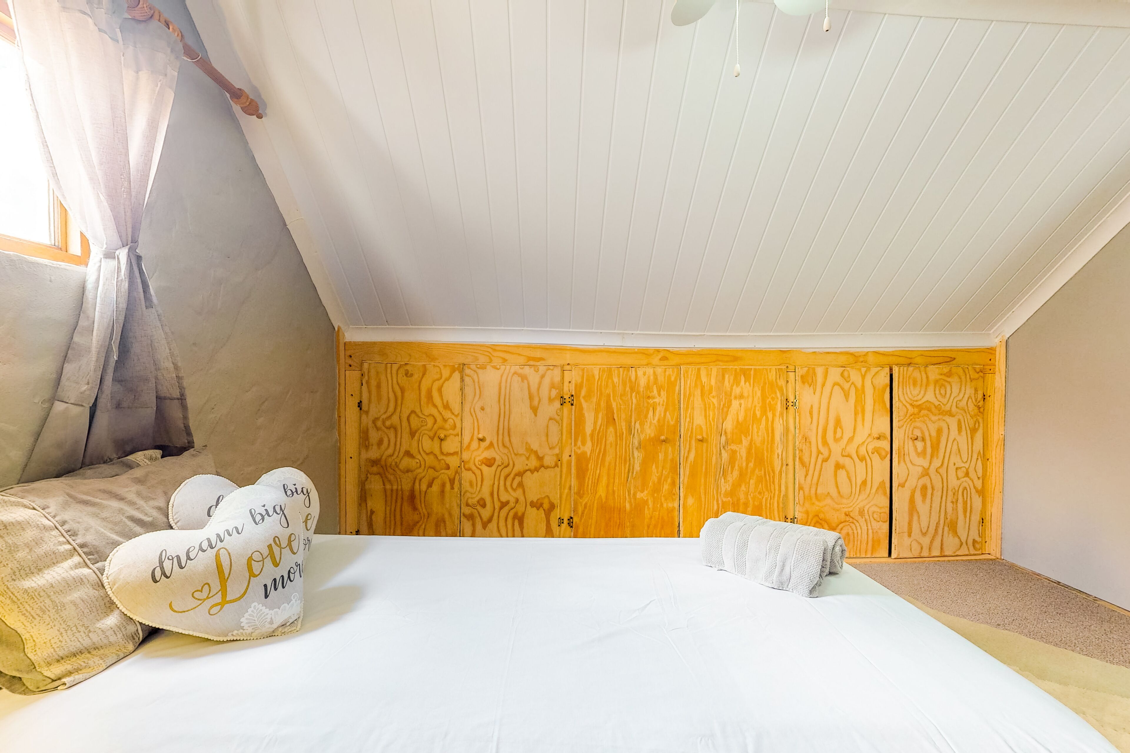Un dormitorio con techo inclinado, paredes de paneles de madera, una cama con ropa blanca y almohadas decorativas.