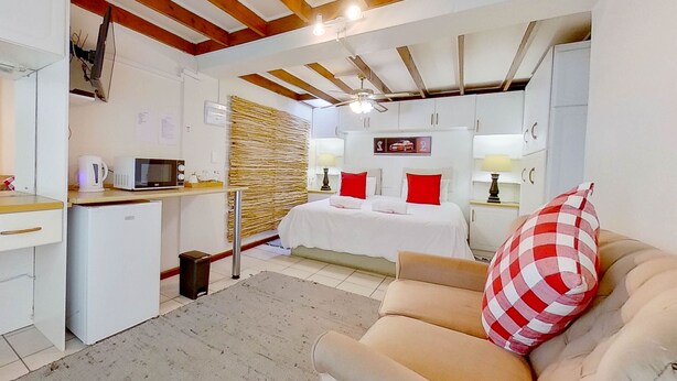A compact room with a kitchenette, a bed with red pillows, a sofa with a checkered pillow, and a ceiling fan. - Executive Couples Suite Strand (Cape Town)