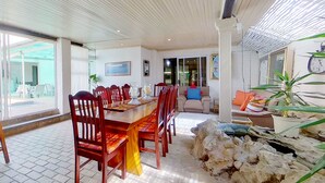 A spacious dining area with a wooden table and chairs, a living area with a sofa and cushions, and a view through large windows. - Executive Couples Suite Strand (Cape Town)
