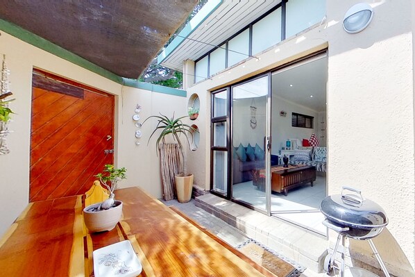 A wooden bench with a potted plant, a grill, and a sliding glass door leading to a living area with a couch and a coffee table.