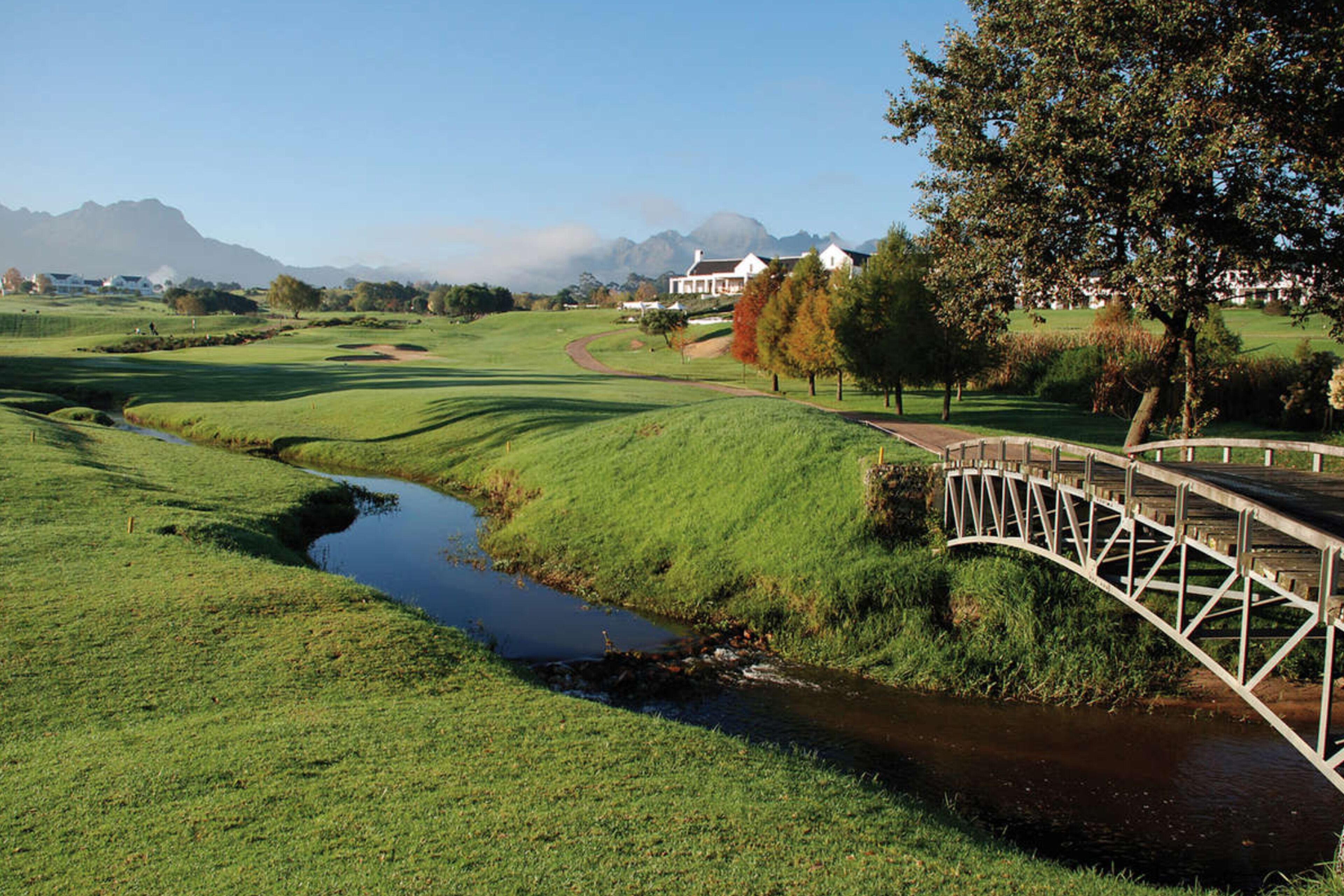 A rural landscape with a bridge over a stream, houses in the background, and a clear sky.