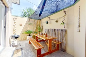 A covered patio with a wooden table and benches, surrounded by potted plants and decorative wall hangings.