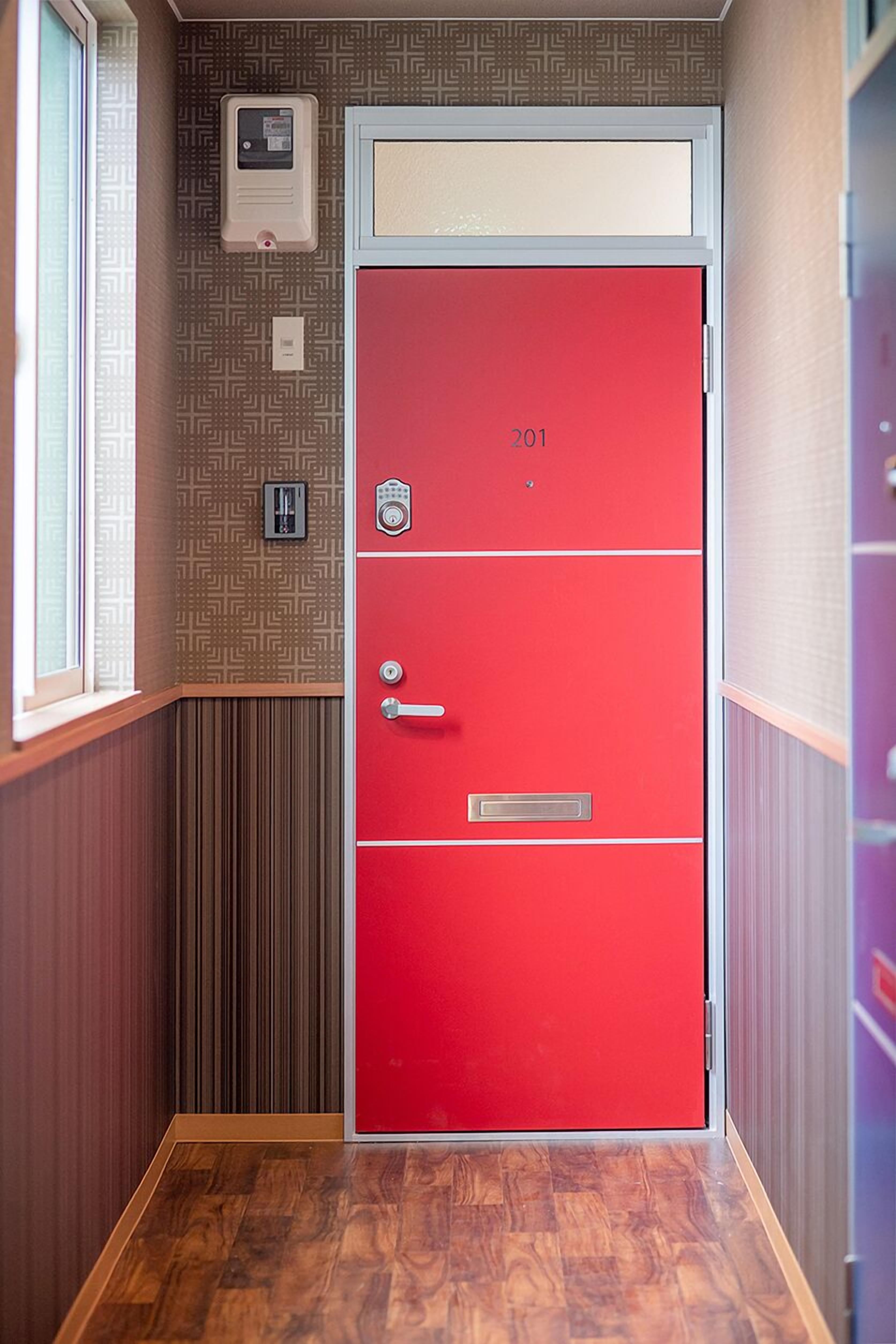 Une porte rouge portant le numéro 201, une fenêtre à gauche et un mur orné de motifs.