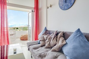 Un salon avec un canapé gris, des coussins bleus, une horloge murale et une vue sur l’extérieur, visible à travers une porte vitrée.