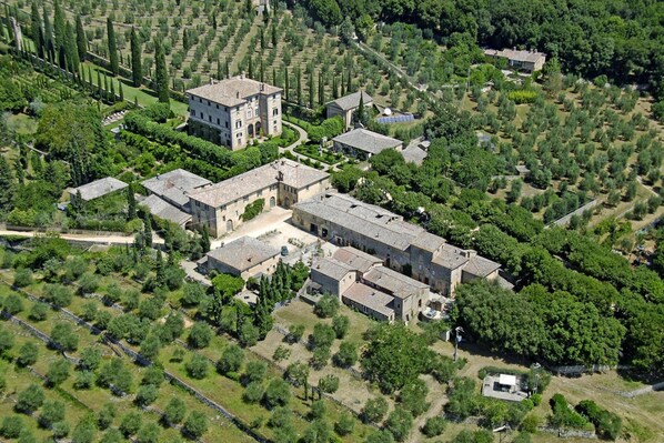 An aerial view of a historic estate with multiple buildings, including a large central structure and smaller surrounding buildings, surrounded by well-maintained gardens and olive groves.