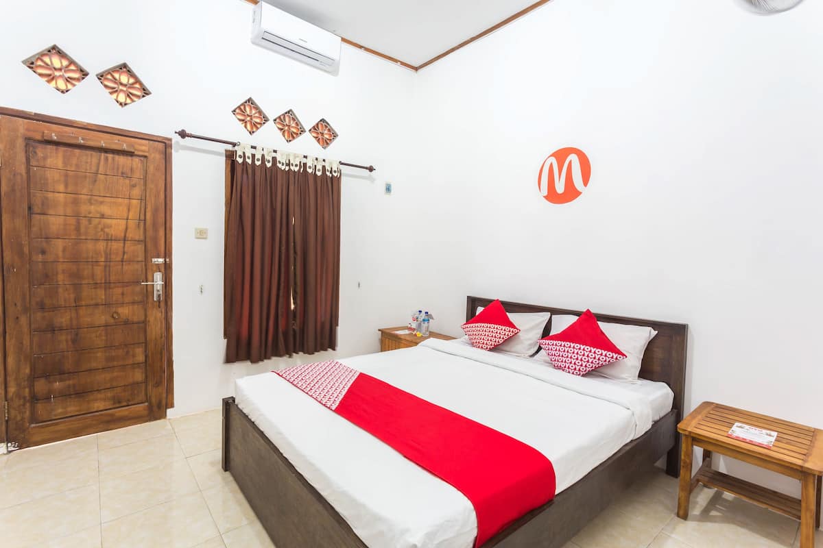 Un dormitorio con puerta de madera, cama con ropa de cama blanca y detalles rojos, mesita de noche con una botella y aire acondicionado instalado en la pared.