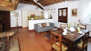 A cozy, rustic room with a wooden dining table set for four, a grey sofa, and a kitchen area in the background. - Charming House in Tuscany with Pool & Views (Sovicille)