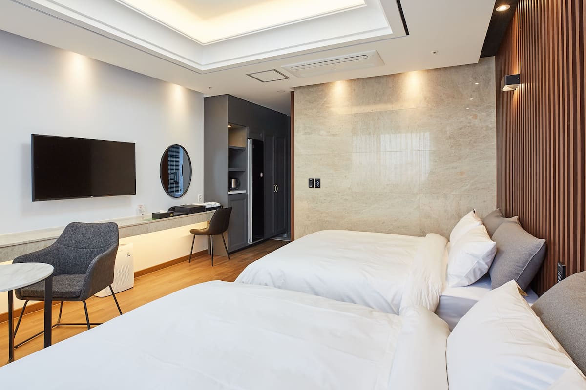 A modern hotel room with a flat-screen TV, a desk with a chair, a bed with white linens, and a wooden panel wall.