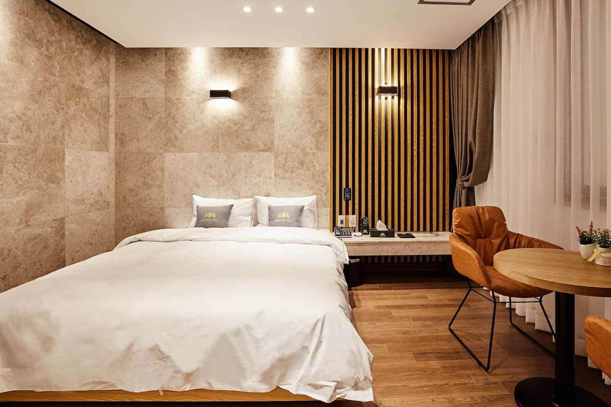 A modern hotel room with a large bed, a wooden side table, a round dining table, and a chair.