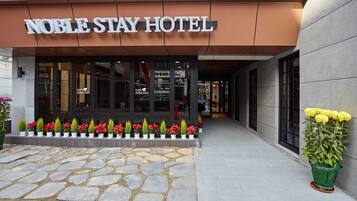 El exterior del Hotel Noble Stay, con un acceso adoquinado, plantas en macetas y una vista despejada de la entrada.