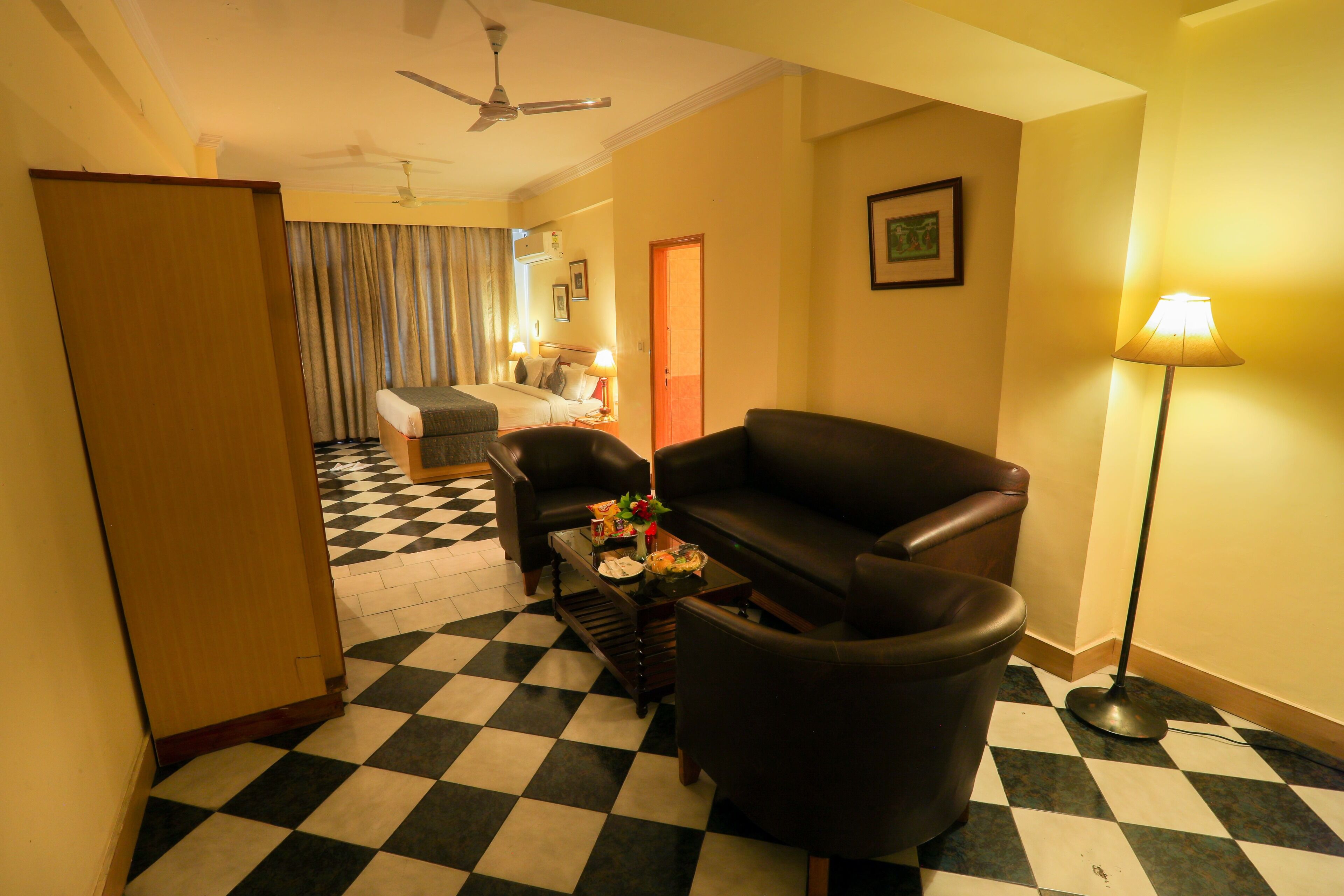 A hotel room with a checkered floor, a sofa, a coffee table, a lamp, and a bed with a headboard.