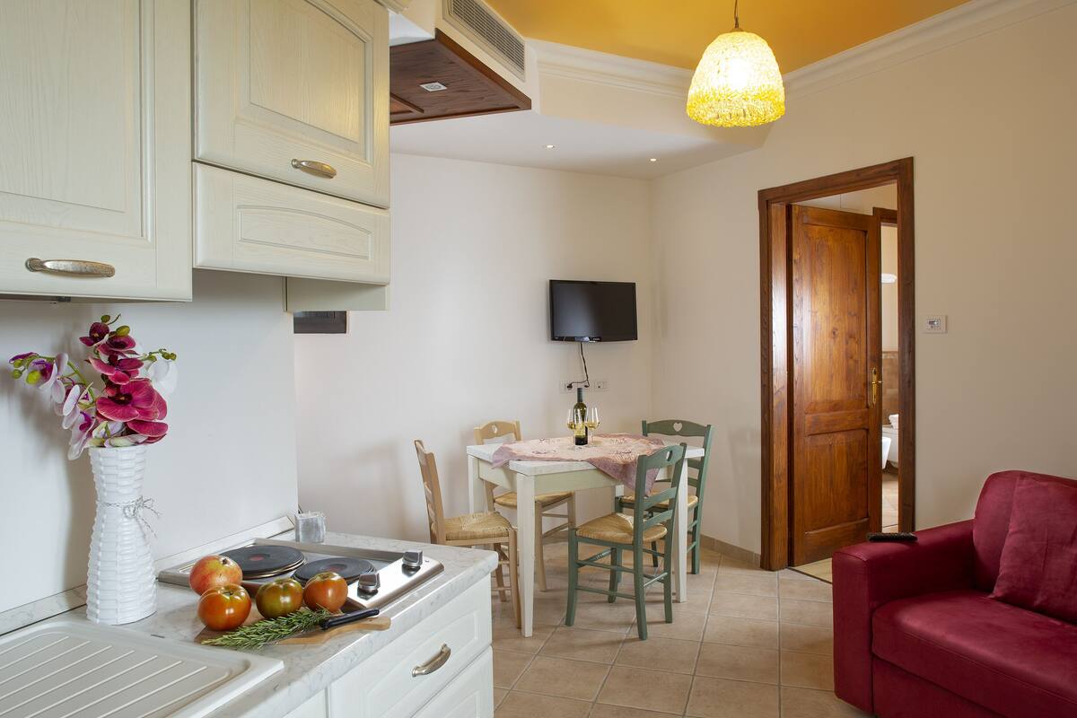 Una cocina con armarios blancos, una mesa de comedor con sillas, un sofá rojo y un televisor en la pared.