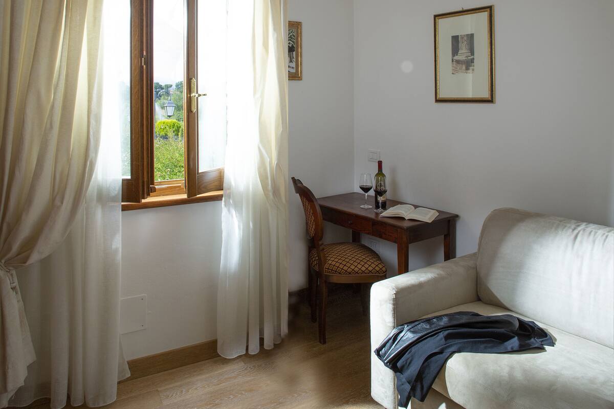 Una habitación con un escritorio de madera, una silla, un sofá blanco y una ventana con cortinas.