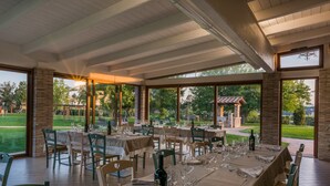 Un espace de restauration en plein air, avec des tables dressées pour le repas, entouré de grandes fenêtres offrant une vue sur un jardin et un bâtiment situé au loin.