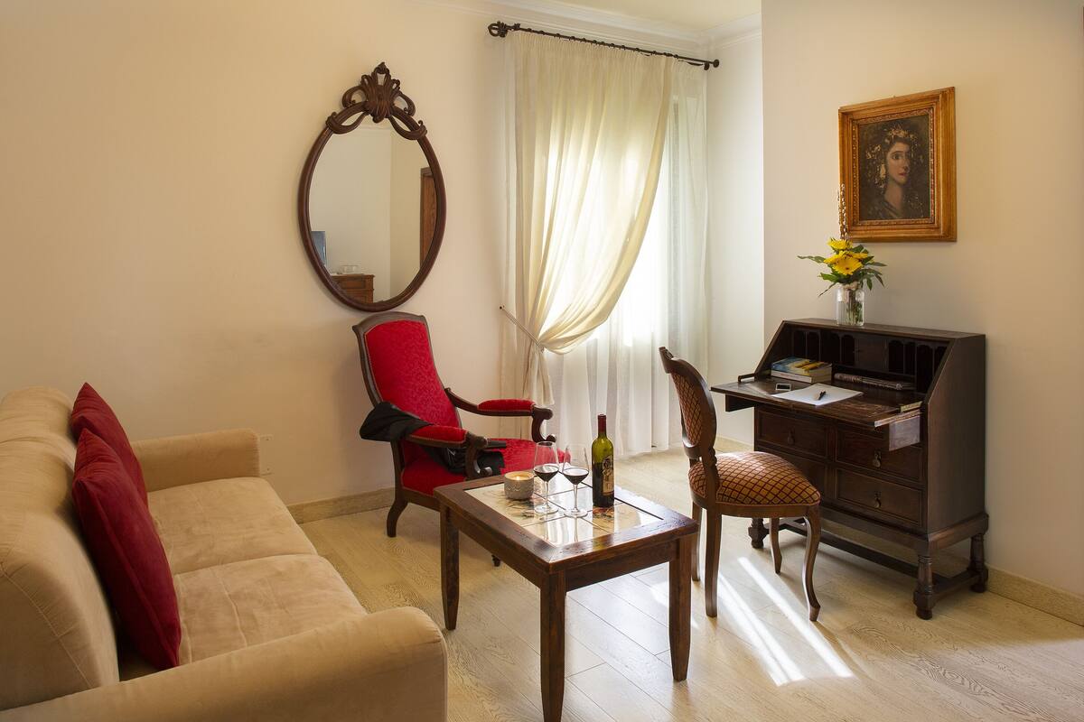Una sala de estar con un sofá color beige, un sillón rojo, una mesa de centro de madera, un escritorio de madera y un espejo en la pared.