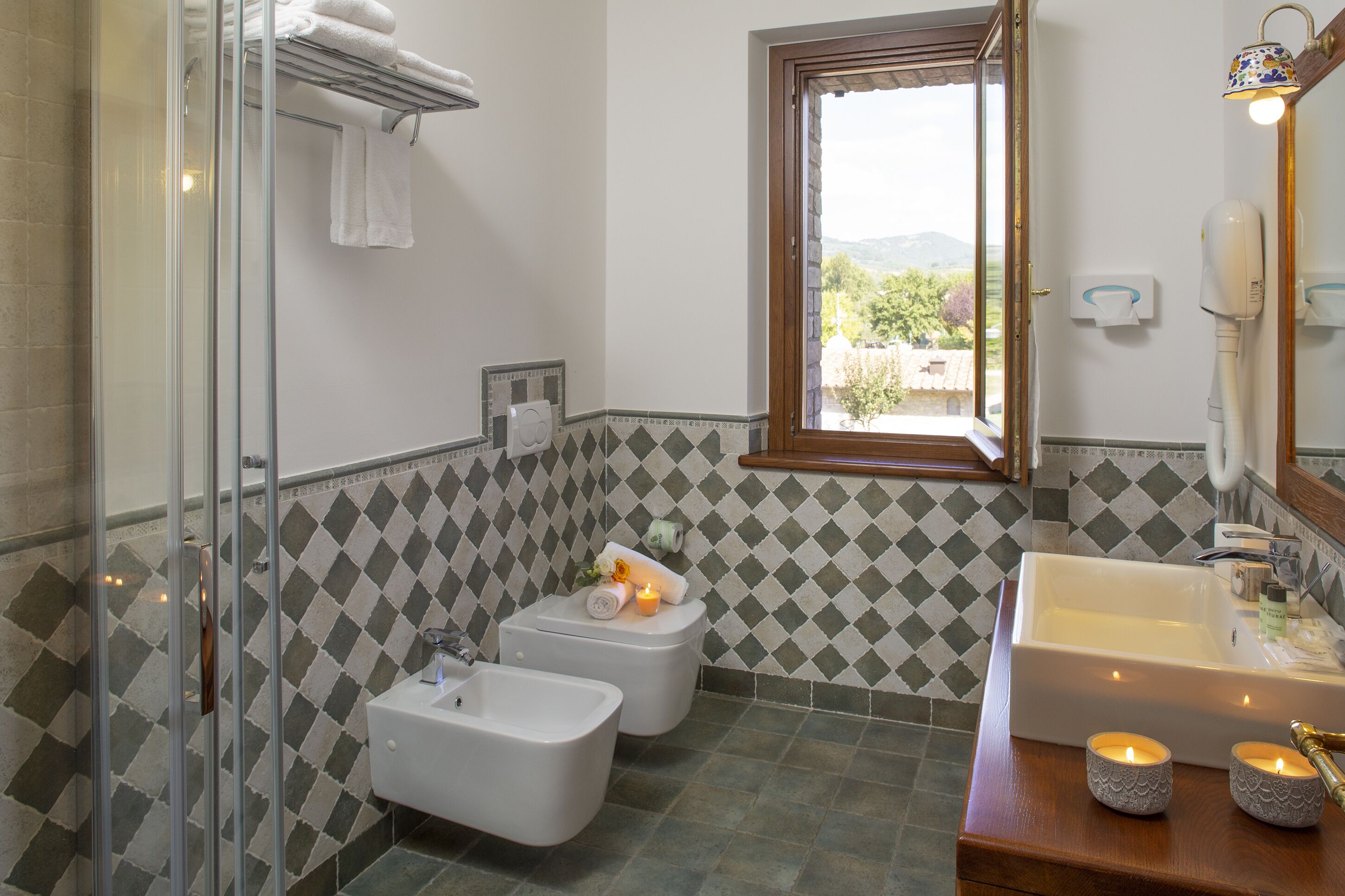 A bathroom with a shower, bidet, sink, and a window with a view of greenery.
