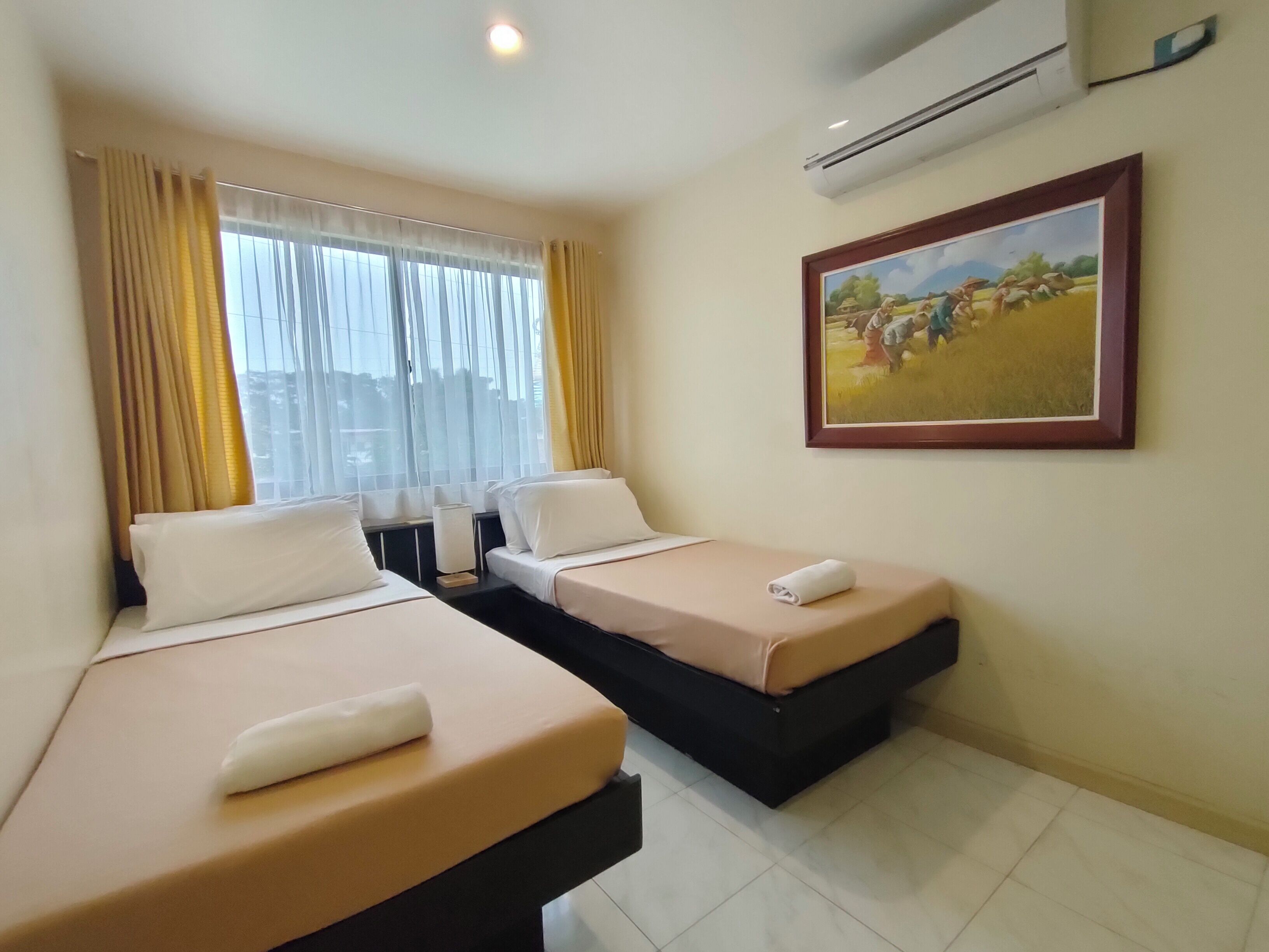 A hotel room with two beds, a window with curtains, an air conditioner, and a framed painting on the wall.