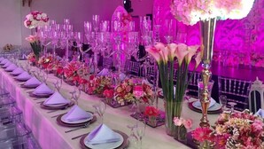 Salão de festas com longas mesas preparadas para um evento formal, com centros de mesa florais e taças altas e transparentes.