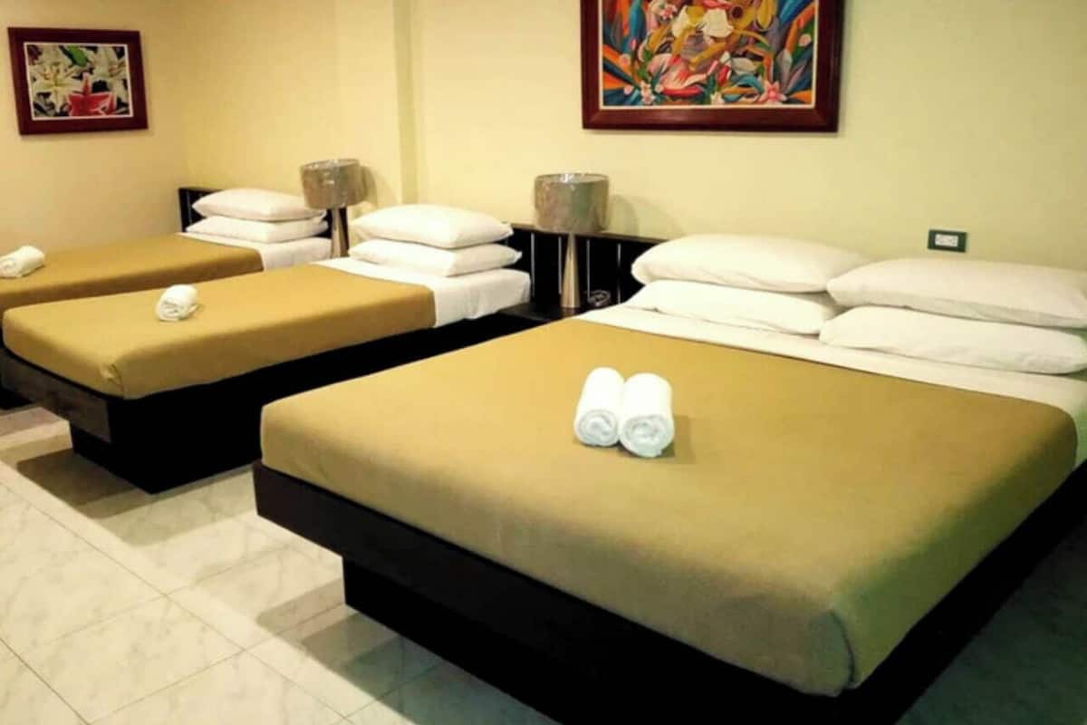 Ein Hotelzimmer mit drei Einzelbetten, jedes mit einer braunen Bettdecke und weißen Kissen. Auf den Nachttischen befinden sich je zwei Wandlampen.