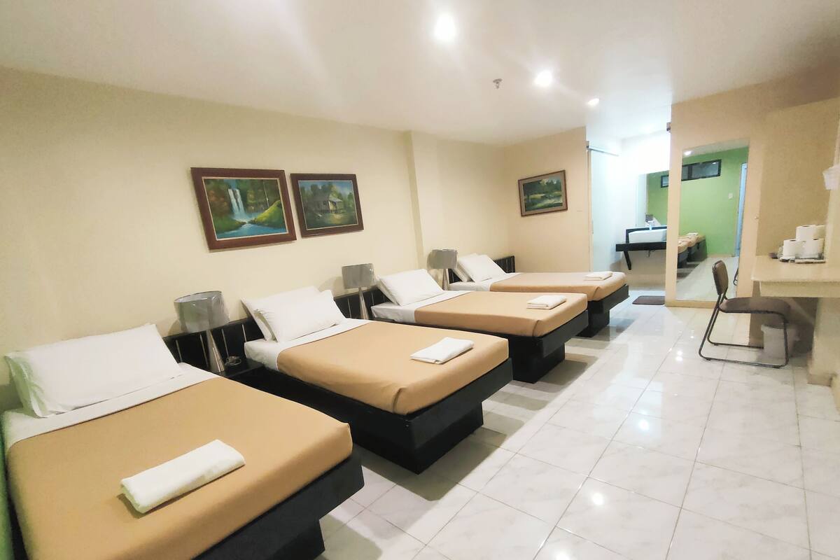 Ein Hotelzimmer mit vier Betten, die nebeneinander aufgereiht sind, jedes mit einem weißen Kopfkissen und einer beige farbenen Decke. Über den Betten hängen zwei Gemälde an der Wand.