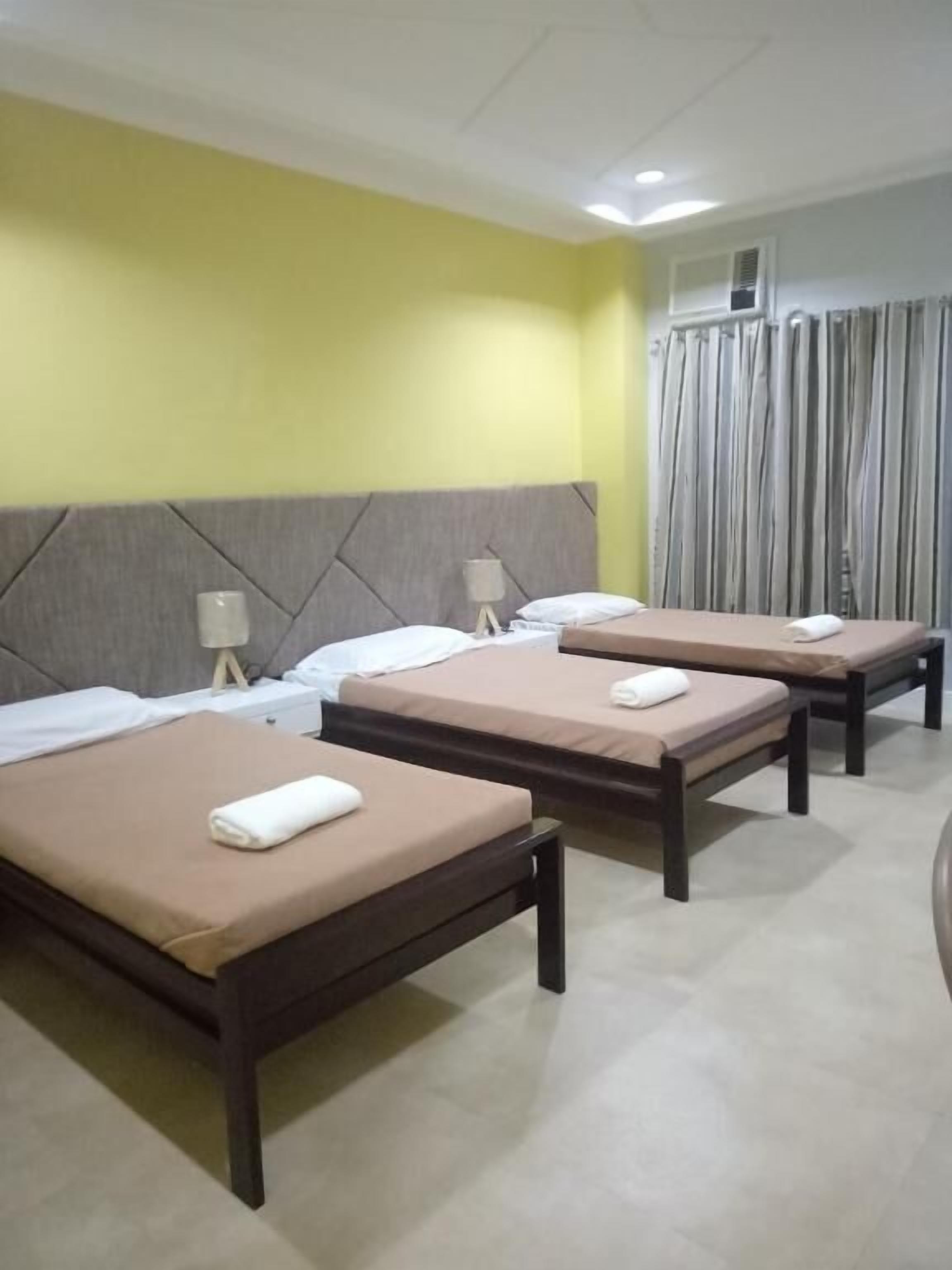 Habitación de hotel con tres camas, cada una con una almohada y una toalla. La habitación tiene una pared amarilla, un cabecero gris y una ventana con cortinas.