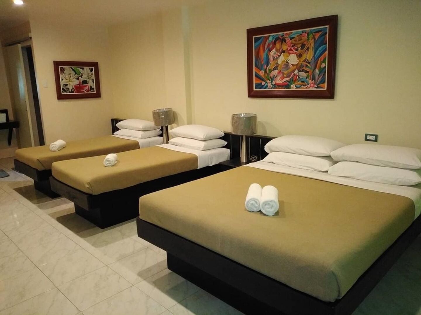 A hotel room with two single beds and a larger bed, each with towels. There are two paintings on the walls.