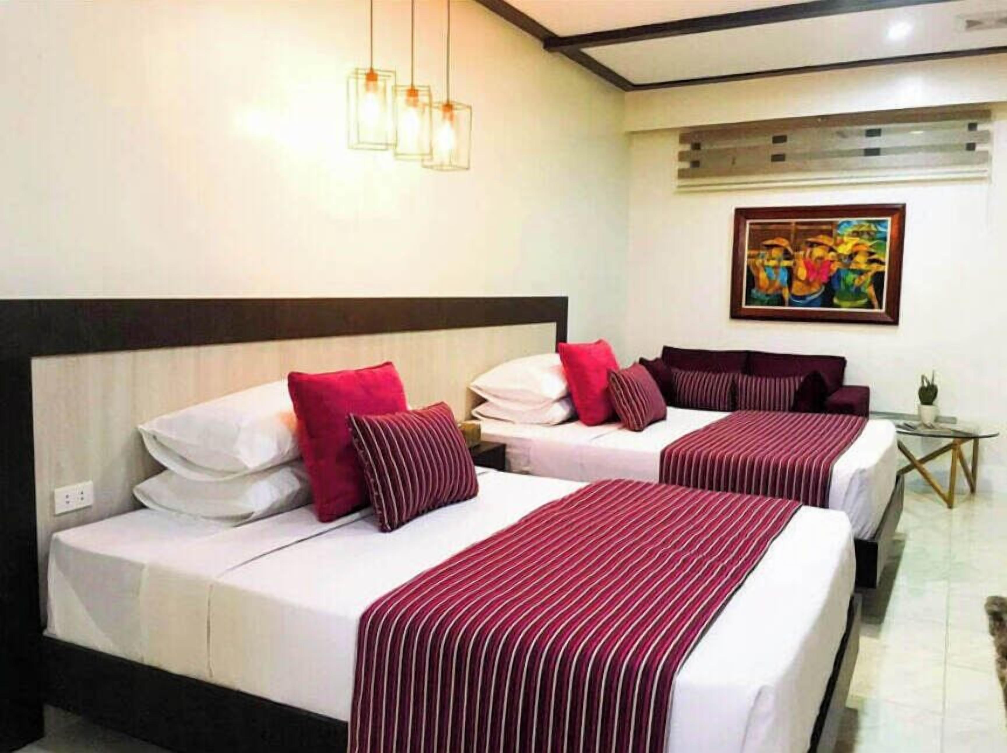 A hotel room with two beds, each with white linens and red striped bedspreads. There are three pendant lights above the beds, and a small table with a plant on the right side.